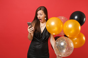 Fototapeta premium Shocked girl in little black dress screaming holding air balloons using mobile phone while celebrating isolated on red background. Valentine's Day Happy New Year birthday mockup holiday party concept.