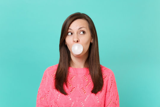 Pretty Young Woman In Knitted Pink Sweater Looking Up, Chewing, Blowing Bubble Gum Balloon Isolated On Blue Turquoise Wall Background. People Sincere Emotions, Lifestyle Concept. Mock Up Copy Space.