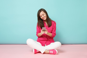 Full length portrait of young fun woman in rose shirt, white pants sit on floor hold cellphone...