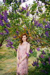 Pretty young brunette girl posing in blooming lilac tree with dark blue flowers outdoor in park. Nice looking tender happy cheerful woman wearing pink stylish feminine dress. Cute female adore nature.