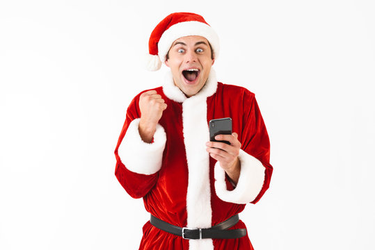 Image of excited man 30s in santa claus costume holding mobile phone, isolated on white background in studio