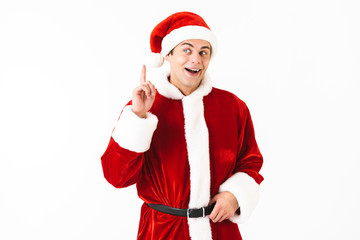 Portrait of young man 30s in santa claus costume and red hat gesturing finger upward, isolated over white background in studio