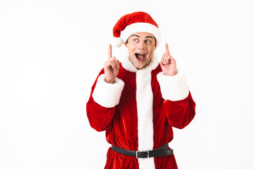 Fototapeta na wymiar Portrait of delighted man 30s in santa claus costume and red hat gesturing fingers upward, isolated over white background in studio