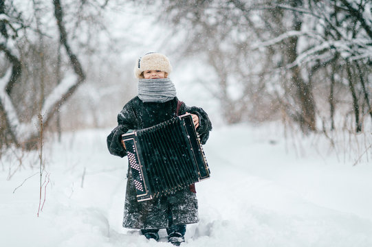 Young Happy Musician. Comic Girl Holding Accordion. Lovely Child In Funny Winter Clothes Outdoor Lifestyle Portrait. Odd Unusual Female Kid Wearing Oversized Adult Padded Jacket. Country Baby Fashion