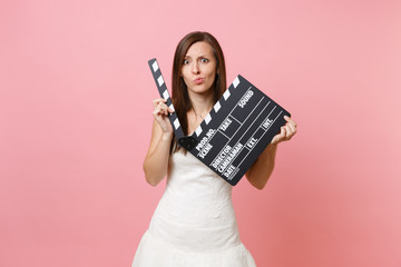 Shocked concerned bride woman in white wedding dress holding classic black film making clapperboard isolated on pastel pink background. Wedding to do list. Organization of celebration. Copy space.