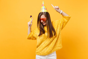 Joyful woman in orange funny glasses birthday party hat with playing pipe rising hands pointing...