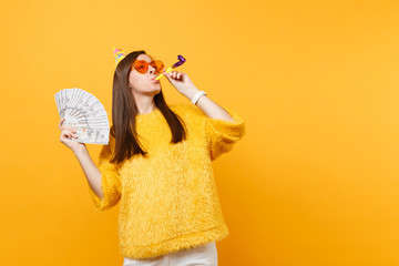 Cheerful young woman in orange heart glasses, birthday hat with playing pipe holding bundle lots of...