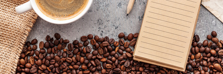 A panoramic photo of a concrete top from above. Opened notebook with brown pages, coffee in a white cup, spilled coffee beans and a jute bag