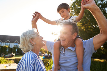 Fototapeta premium Grandparents Giving Grandson Ride On Shoulders In Summer Park Against Flaring Sun