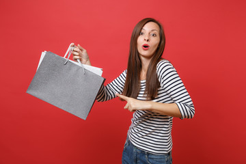 Fototapeta na wymiar Amazed young woman pointing index finger on packages bags with purchases after shopping in hands isolated on bright red wall background. People sincere emotions, lifestyle concept. Mock up copy space.