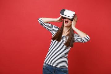 Excited young girl in striped clothes, virtual reality glasses keeping mouth wide open, putting...