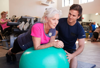 Fototapeta na wymiar Senior Woman Exercising On Swiss Ball Being Encouraged By Personal Trainer In Gym
