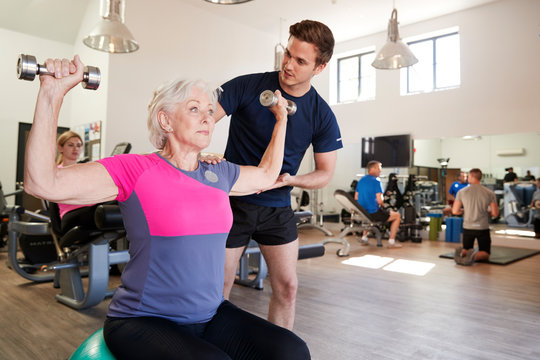 Senior Woman Exercising On Swiss Ball With Weights Being Encouraged By Personal Trainer In Gym