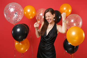 Happy girl in black dress celebrating, holding glass of champagne, clenching fist like winner on bright red background air balloons. Women's Day, Happy New Year, birthday mockup holiday party concept.