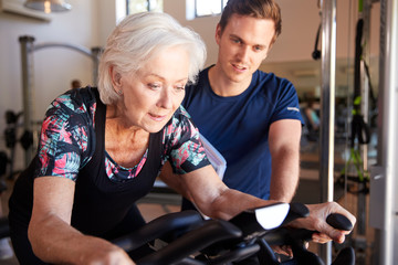 Fototapeta na wymiar Senior Woman Exercising On Cycling Machine Being Encouraged By Male Personal Trainer In Gym