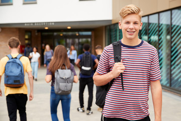 Portrait Of Smiling Male High School Student Outside College Building With Other Teenage Students...