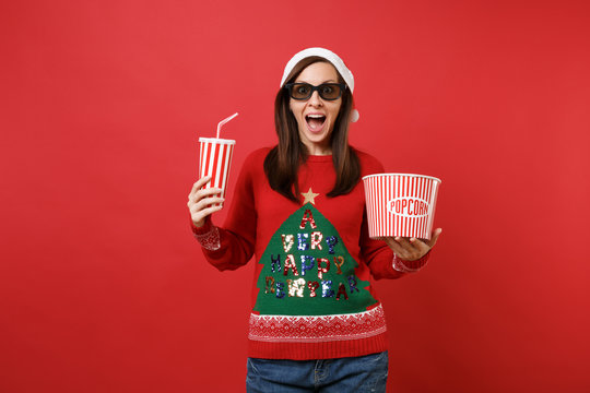 Excited young Santa girl in 3d imax glasses watching movie film holding popcorn, cup of soda isolated on red wall background. Happy New Year 2019 celebration holiday party concept. Mock up copy space.