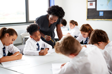 Female High School Tutor Helping Students Wearing Uniform Seated Around Tables In Lesson