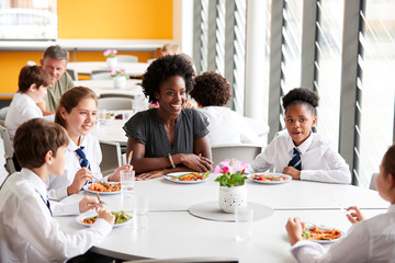 Female Teacher With Group Of High School Students Wearing Uniform Sitting Around Table And Eating...