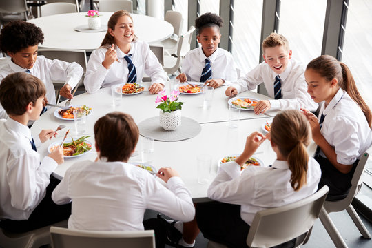 Group Of High School Students Wearing Uniform Sitting Around Table And Eating Lunch In Cafeteria