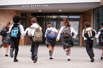Group Of High School Students Wearing Uniform Running Into School Building At Beginning Of Class