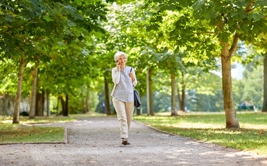 retirement, communication and old people concept - happy senior woman walking along summer park and...