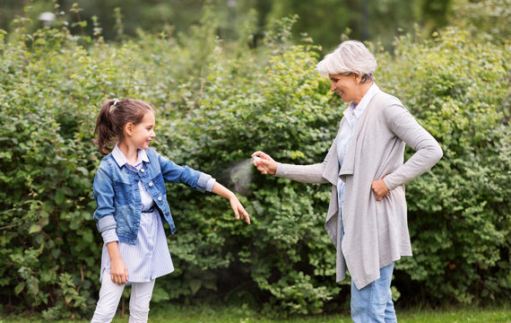 Family, Leisure And People Concept - Happy Grandmother And Granddaughter Applying Insect Repellent Spray At Summer Park