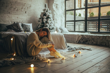 Young woman sitting near bed and hugging himself on background of Christmas tree, glowing lightbulbs and window.