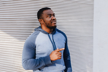 Portrait of a black man in a blue sports sweater on the background a striped white wall. He looks up, shows a finger. Relaxed state, city photo