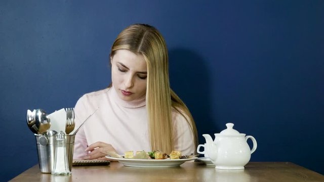 Young blonde girl eating waffles for breakfast, drinking tea and taking notes in notepad. 4K