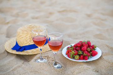 straw hat, two glasses of wine and a plate of strawberries on a sandy beach, close-up.