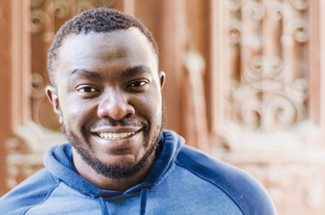Fototapeta premium Portrait of a cheerful positive black guy close-up in a sports sweater on the background of an urban house. Race issues. City photo, space. Youthful culture.