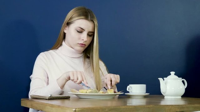 Young blonde girl eating waffles for breakfast, drinking tea and taking notes in notepad. 4K