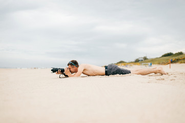 Young traveler man lying on beach near sea on vacation and making photos of nature. Topless handsome barefoot man with camera and lense spying people from afar. Time for hobby. Boy resting on sand