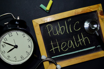 The words Public Health handwriting on chalkboard on top view. Alarm clock, stethoscope on black...