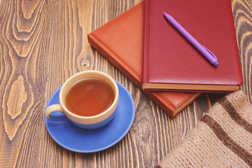 Blue tea cup with notebooks, a pen and a scarf on wooden table.
