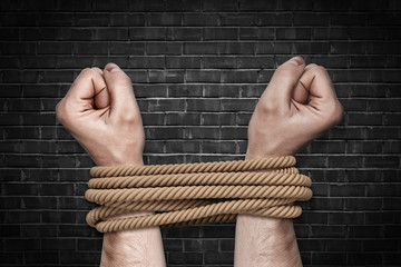 Two bound strong male arms struggling to break a strong natural rope on a black brick background.