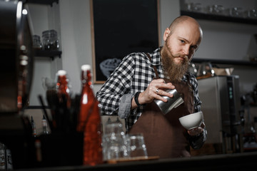 Brutal young barista in an apron makes coffee at the bar in a modern cafe