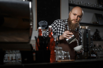Brutal young barista in an apron makes coffee at the bar in a modern cafe