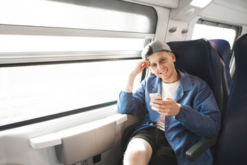 Portrait of a smiling boy in casual clothes sitting in a train near the window with a smartphone in his hands, looking into the camera and smiling. Happy train trainer smiles.
