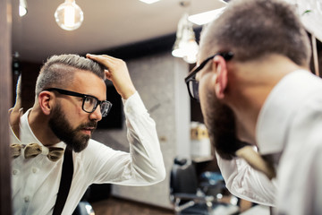 Hipster man client with glasses looking in the mirror in barber shop.