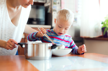 A handicapped down syndrome boy pouring soup in a plate indoors, lunch time.