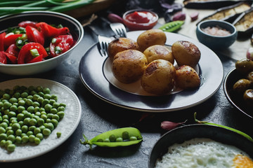 Delicious dinner table for two with fried potatoes, organic tomatoes salad, olives, green onion, eggplants, eggs on dark stone background.