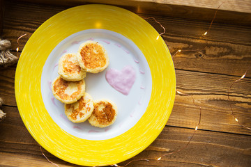 cheese pancakes on a yellow plate, a wooden tray, lights, a decoration in the form of a heart.