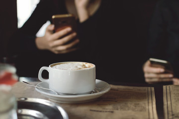 A cup of cappuccino coffee on a table in a cafe. People drink coffee in a cafe and use mobile phones. Close-up, selective focus.