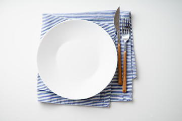 White empty plate with knife and fork on striped blue and white textile napkin on white background