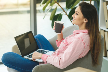 Enjoying working time at home. Beautiful young smiling casual woman working on laptop and drinking coffee while sitting in a big comfortable chair at home.