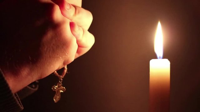 Man clasping hands with cross, praying near a burning candle