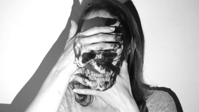 Skull projection on womans face. She covers herself as if to hide from death. Black and white. Skull image is in the public domain.