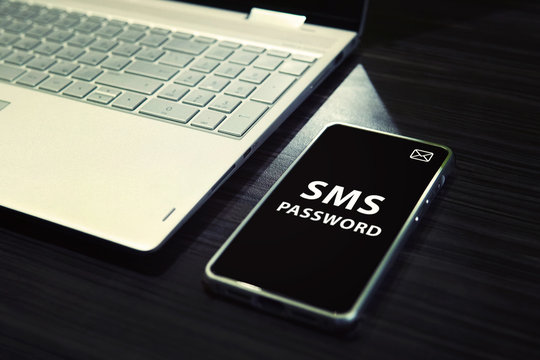 Table With Open Laptop And Black-screened Smartphone With White Written Words SMS Password On Its Display. Concept Of Linking The Work Of Computer And Mobile Via Sms Password Entering. One Time Code.